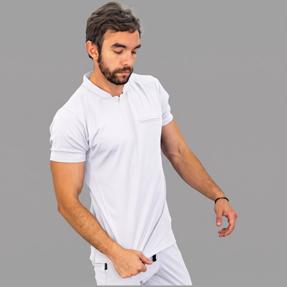 scrubs-blanco-mirada-abajo.jpg: "Modelo mirando hacia abajo con la filipina quirúrgica Est. 910 blanca, mostrando su corte slim fit y el bolsillo del pecho con cierre."